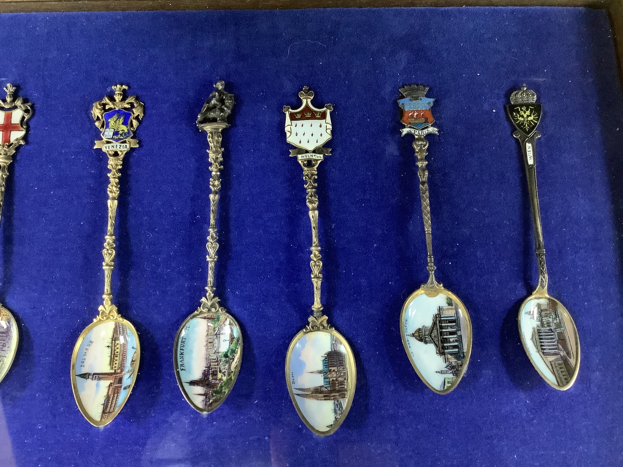 A collection of sixty four 800 standard silver (many gilt) and enamel souvenir spoons, early 20th century, in eight display cases, Including views of London, Germany, Italy, Austria, Spain etc.